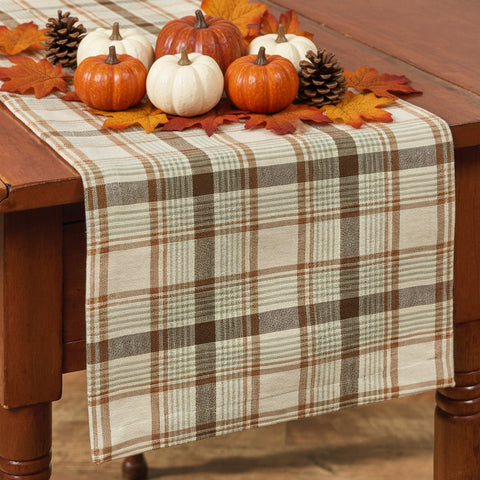 Toffee & Elm Plaid Table Runner 36" draped over a wooden dining table, featuring a cream, brown, and tan checkered pattern with Fall pumpkin leaves and pinecone accents.