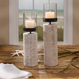 Two rustic white-washed Distressed White Log Votive Candle Holders with black metal rings and clear glass cylinder candle holders, set on a wooden table with a beach view in the background.