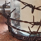 A close-up of Barbed Wire Rustic Pillar Candle Holder featuring dark brown twisted barbed wire wrapped around a clear glass jar filled with a white wax melt or candle.