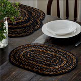 Blackstone Farm Braided Oval Placemat Set of 6 –10x15 – Black and Tan Jute on a dark wood dining table, styled with a white bowl and plate, silverware, a stack of matching placemats, and plant arrangement with a wooden chair.