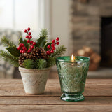 Fleur de Lys Green Tumbler Hand-Poured Soy Scented Candle is a lit green glass candle holder with an embossed floral pattern sits on a rustic wooden table next to a small potted holiday plant with red berries, pinecones, and holly leaves, with a softly blurred fireplace in the background.