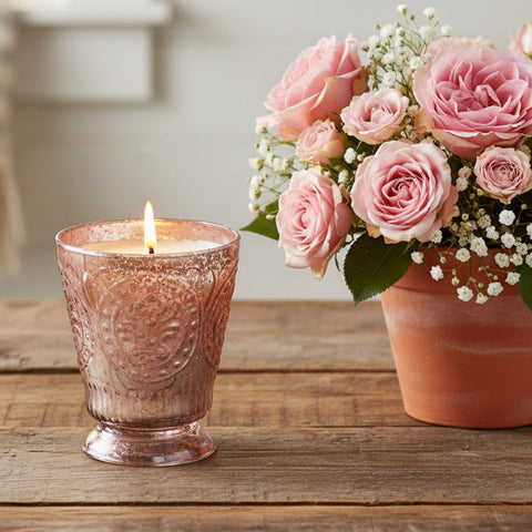 Fleur de Lys Pink Tumbler Hand-Poured Soy Scented Candle in a textured, rose pink-silver-toned glass holder sits on a rustic wooden table next to a terracotta pot of pink roses and baby's breath.