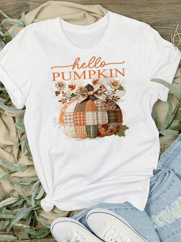 Hello Pumpkin Patchwork Fall T-Shirt - A white graphic t-shirt featuring a 'hello PUMPKIN' design with a plaid patchwork pumpkin, styled in a flat-lay with beige fabric, green leaves, light blue jeans, and white sneakers.