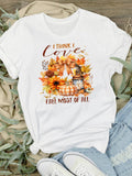 I Think I Love Fall Most Of All T-Shirt - A white short-sleeve graphic T-shirt laid flat on a wood surface with beige fabric, greenery, jeans, and white sneakers around it. The shirt has a fall-themed print with the text 'I THINK I love FALL MOST OF ALL' and an autumn floral design in orange, brown, yellow, and beige tones.