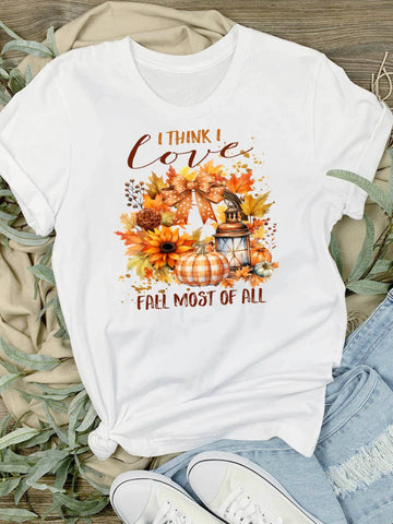 I Think I Love Fall Most Of All T-Shirt - A white short-sleeve graphic T-shirt laid flat on a wood surface with beige fabric, greenery, jeans, and white sneakers around it. The shirt has a fall-themed print with the text 'I THINK I love FALL MOST OF ALL' and an autumn floral design in orange, brown, yellow, and beige tones.
