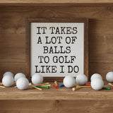 A square framed decorative sign with a rustic brown wood frame and a white face on a wood shelf with golf balls and golf tees. The sign features the humorous text 'IT TAKES A LOT OF BALLS TO GOLF LIKE I DO'.