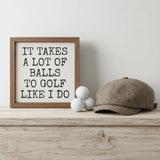 A square framed decorative sign with a rustic brown wood frame and a white face. Golf balls and golf hat on a wood shelf.  The sign features the humorous text 'IT TAKES A LOT OF BALLS TO GOLF LIKE I DO' in a black, hand-drawn serif font.