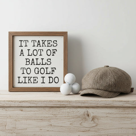 A square framed decorative sign with a rustic brown wood frame and a white face. Golf balls and golf hat on a wood shelf.  The sign features the humorous text 'IT TAKES A LOT OF BALLS TO GOLF LIKE I DO' in a black, hand-drawn serif font.