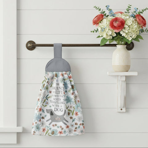 A fabric pet memorial towel hangs from a dark metal wall rod. The towel has a white background with a floral pattern in muted blue, green, and orange-brown tones, featuring a gray dog silhouette and the printed text 'THE JOURNEY OF LIFE WE MEET WHEN TRAVELING WITH A DOG'.