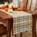 Sage & Mustard Plaid Chindi Table Runners with a plaid pattern in mustard, white, sage green, and brown, styled on a wooden dining table with autumn decorations including pumpkins, candles, and fall leaves.