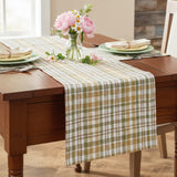 Sage & Mustard Plaid Table Runners in sage green, light mustard, putty brown, and white is draped across a wooden dining table set with plates, napkins, and a vase of Spring pink peony and daisy flowers.