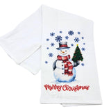 A white kitchen tea towel featuring a festive Christmas design with a watercolor-style snowman wearing a red and white striped scarf, a small green Christmas tree, blue snowflakes, and the words 'Merry Christmas' in red script.