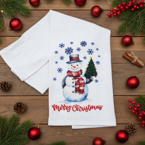 A white kitchen flour sack tea towel on a wood table featuring a festive Christmas design with a snowman wearing a red and white striped scarf, a small green Christmas tree, blue snowflakes, and the words 'Merry Christmas' in red script.