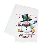 A white kitchen tea towel featuring a festive illustration of a snowman wearing a black top hat and a red patterned scarf, holding a glass of red wine. The text 'WINE is cheaper than THERAPY!' is printed below in red and black.
