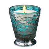 Fleur de Lys Turquoise Tumbler Hand-Poured Soy Scented Candle  is a teal-blue embossed glass candle holder with a lit white candle inside, set against a plain white background.